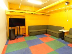 黄色い壁の部屋に大型モニターとソファーが置かれた学生ラウンジ