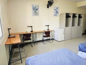 3台のワードローブ、冷蔵庫、勉強机、壁付け扇風機のある3人部屋