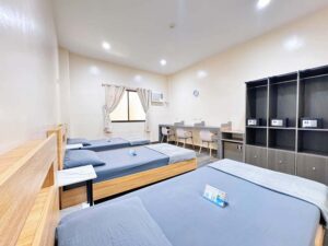 壁際に3人用の机と椅子、冷蔵庫、ワードローブが並ぶ3人部屋