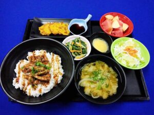 照り焼き丼、味噌汁、野菜炒め、サラダ、フルーツが並ぶ食事