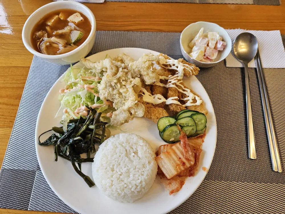 チキンカツ、天ぷら、サラダ、野菜炒め、ご飯に韓国風スープとフルーツサラダが添えられたランチ