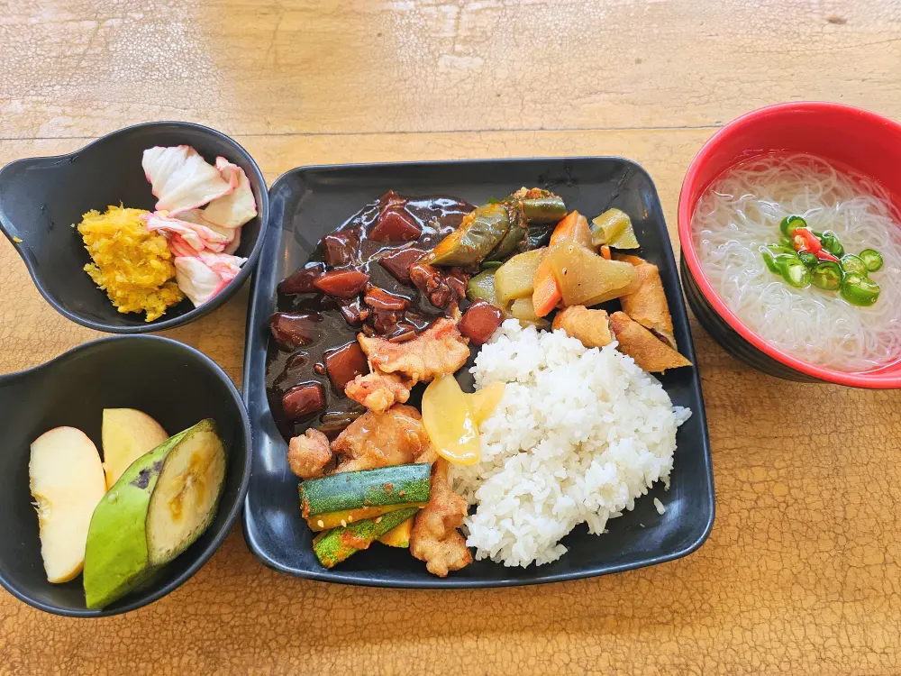 ルンピア、豚肉のフライ、チキンシチューが盛られた皿に、サラダ、麺、フルーツが添えられた昼食