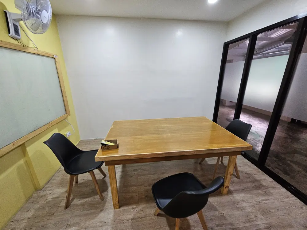 大きな木製テーブルと3脚の椅子が置かれ、ホワイドボードと扇風機が壁に設置されたグループ用教室
