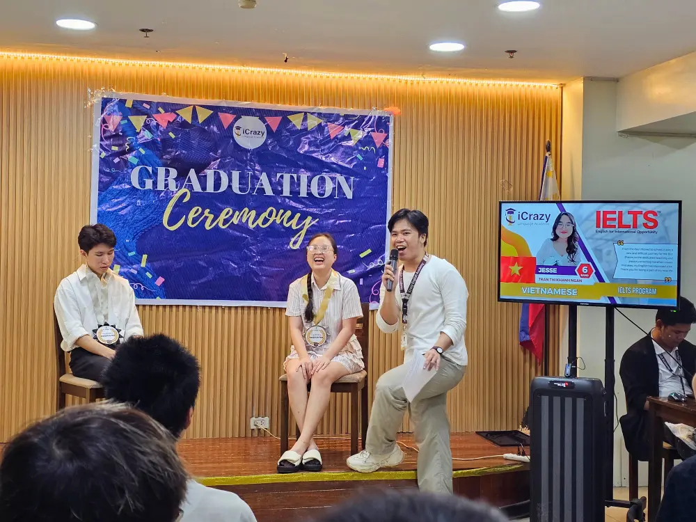 ICLの卒業式で笑顔で司会をしている講師とステージ上の2名の卒業生