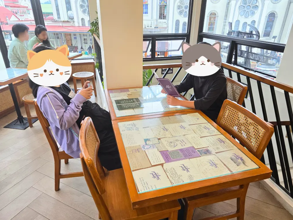 大きな窓からバギオ大聖堂が見える、木製テーブルと籐張りの椅子が置かれたGood Shepherd Caféの2階席