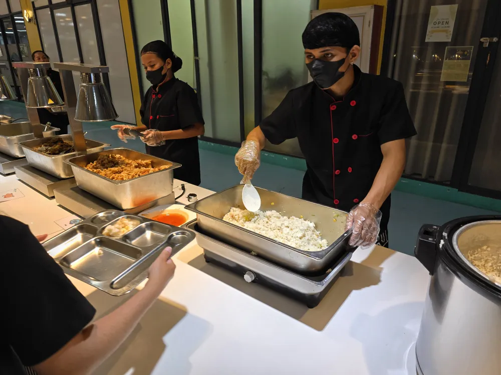 フィリピンのセブ・マクタン島にあるB'Cebuのランチタイムに、ブッフェカウンターでご飯や料理を取り分けるスタッフと食事を選ぶ学生