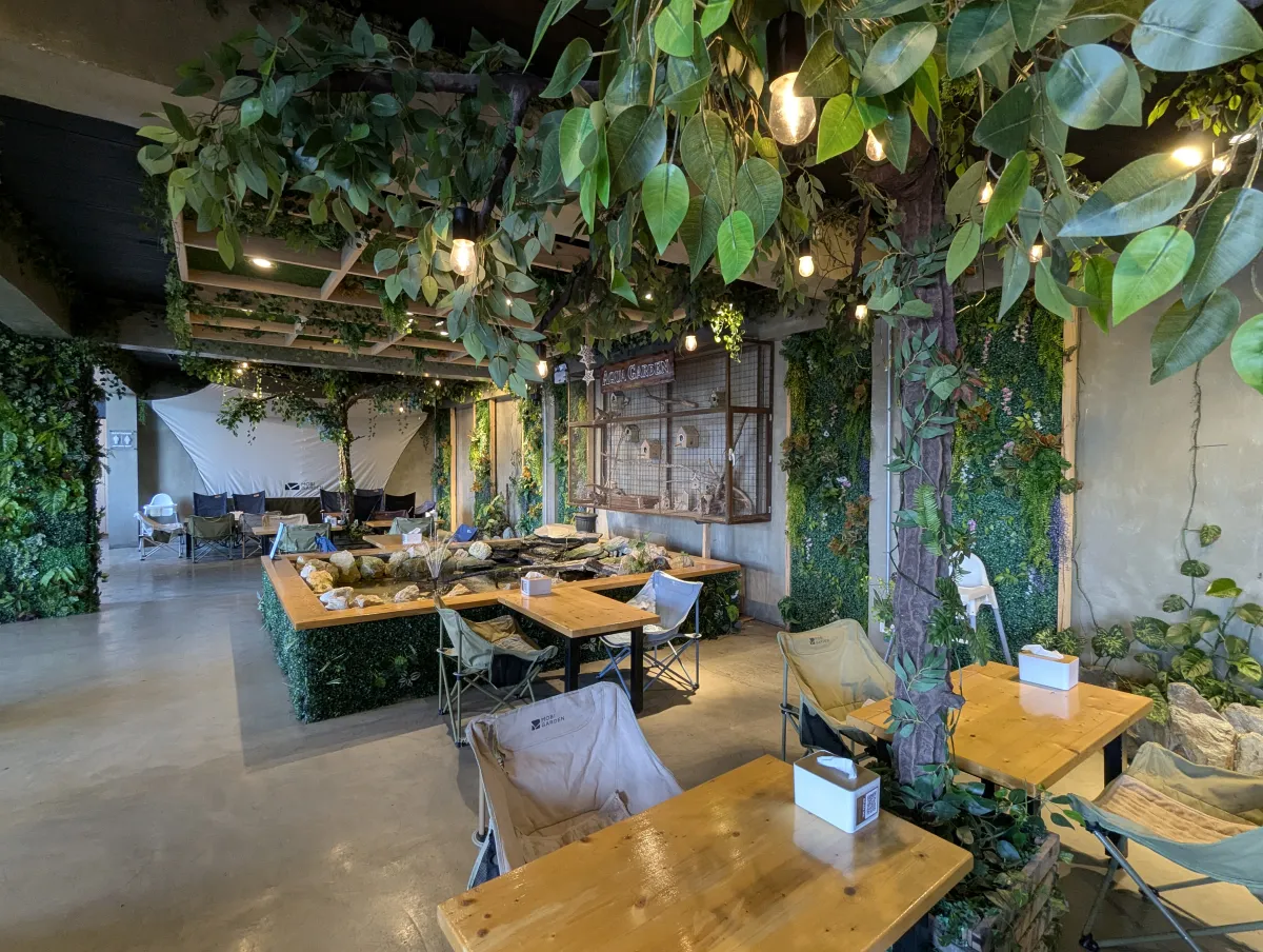 MONOL Main CampusのAqua Garden Café、緑に囲まれた店内に並ぶ木製テーブルと椅子、魚が泳ぐ四角い池