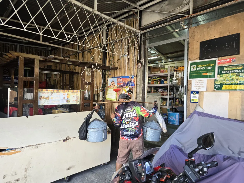 サリサリストアFelomina’s Storeの前で、天秤棒の両端に金属製の容器を下げてフィリピンの伝統菓子プトを売る行商人