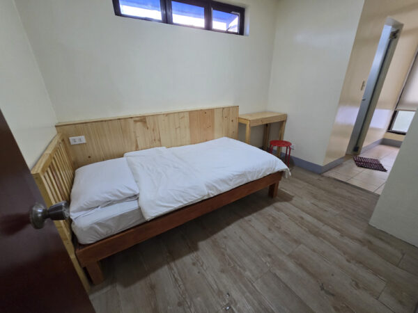 入口側から見た、木製ベッドと勉強机、壁上部の小窓、奥にバスルームがある1人部屋Ｂ