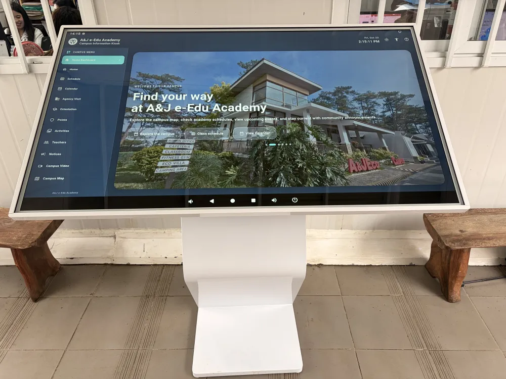 バギオのA&J ECO Campusに導入された大型タッチパネル式のKIOSK情報端末。画面には学校施設の写真とキャンパス案内が表示され、左側には授業スケジュール、カレンダー、アクティビティ、講師情報、キャンパスマップなどのメニューが並んでいる。