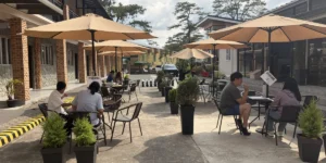 フィリピン・バギオのA&J ECO Campusで始まった屋外マンツーマン授業。学生寮とダイニングホールの間にベージュ色のパラソルとテーブルが並び、学生と講師が向かい合って授業を受けている様子。周囲には植木鉢が置かれ、奥には松の木が見える。