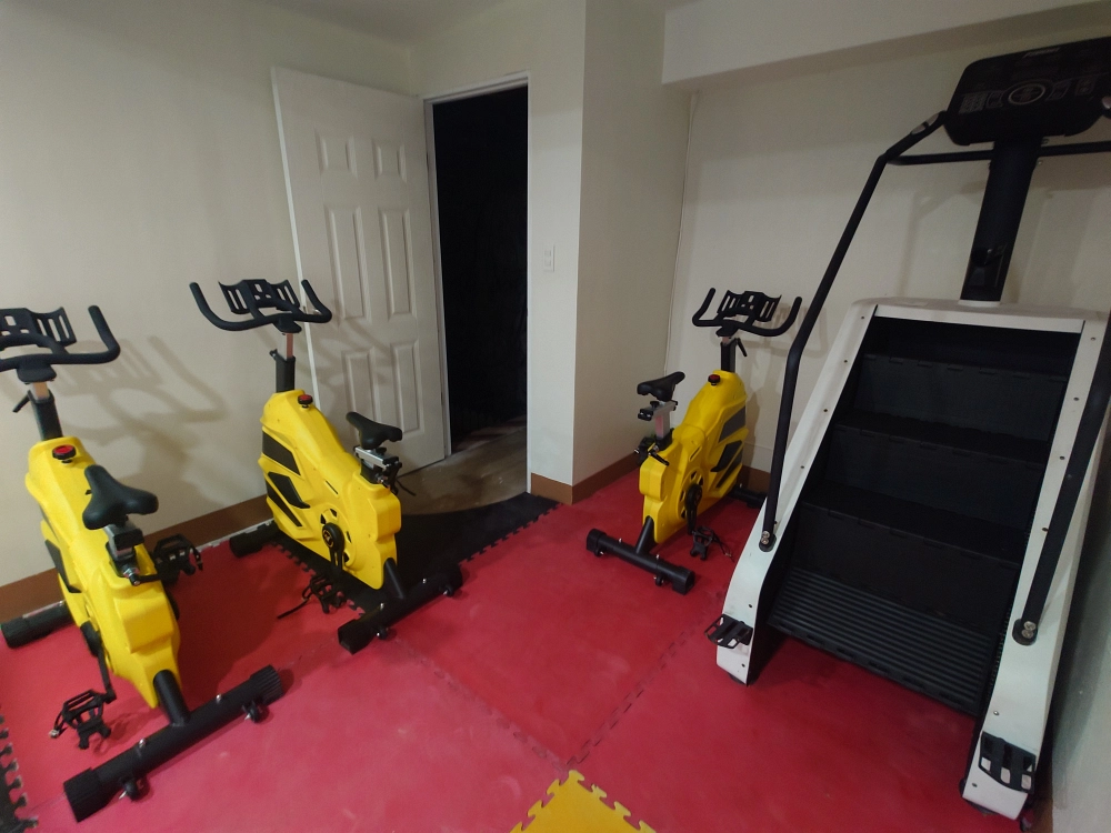フィリピンのバギオにあるMONOL Sparta Campusの校内ジムに設置された3台の黄色いスピンバイクと、階段昇降運動用のクライムミル。赤い床マットの上に有酸素運動用のトレーニングマシンが並んでいる